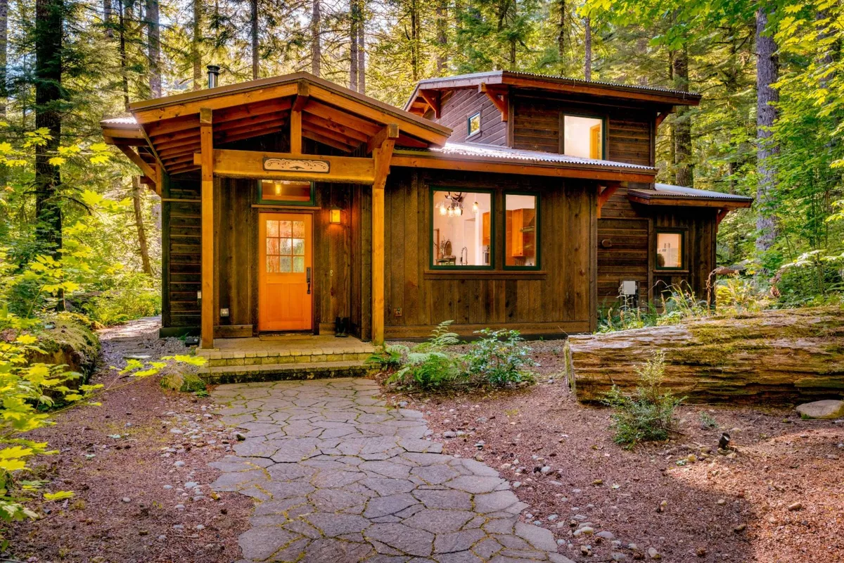 The image showcases the inviting wooden front door of a custom-built cabin, a charming stone pathway leading through vibrant, lush greenery, and the warm wood exterior nestled among towering trees. The scene radiates tranquility, hinting at peaceful mornings, evenings spent outdoors, and a deep connection with nature.