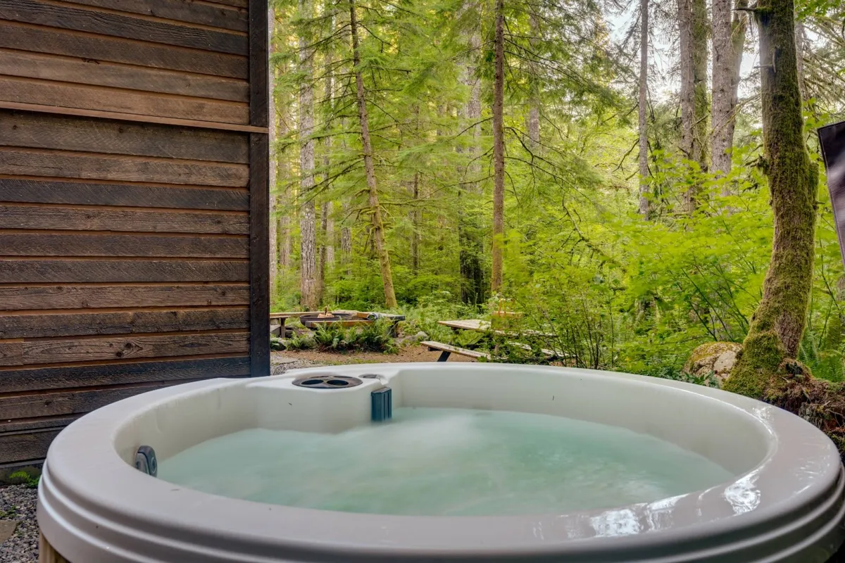 This image shows a tranquil outdoor setting with a hot tub nestled among tall trees, accompanied by picnic tables and surrounded by greenery. The environment is serene, inviting relaxation and immersion in woodland nature.