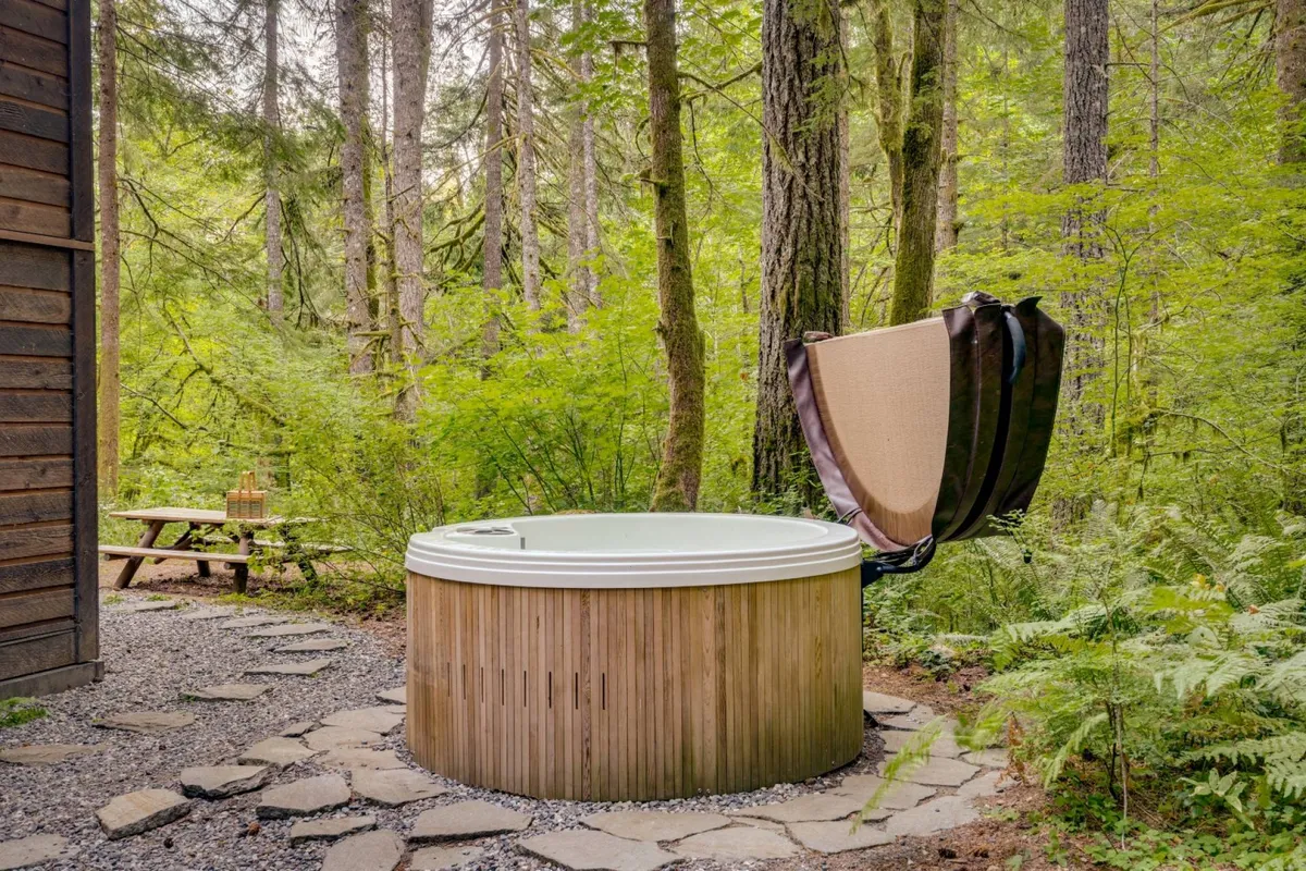 This exterior image features a hot tub tucked among tall trees, with a picnic table nearby and a lush green backdrop. The secluded setting offers a private, immersive experience surrounded by nature, perfect for tranquil relaxation or soaking under the stars.