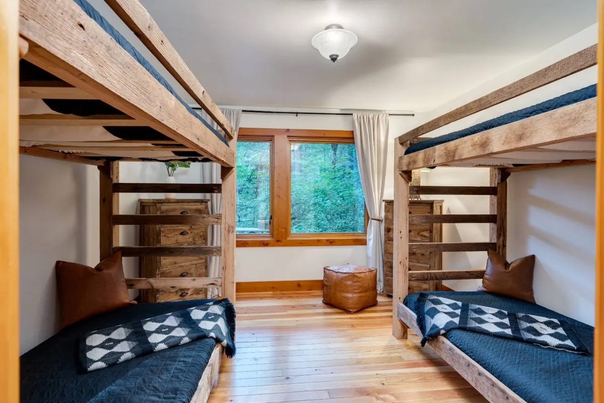 A bunk room with four plush bunk beds, large windows letting in natural light, wooden furniture, and decorative pillows. The mood is friendly and relaxed, with a sense of playful comfort perfect for kids or friends sharing the space.