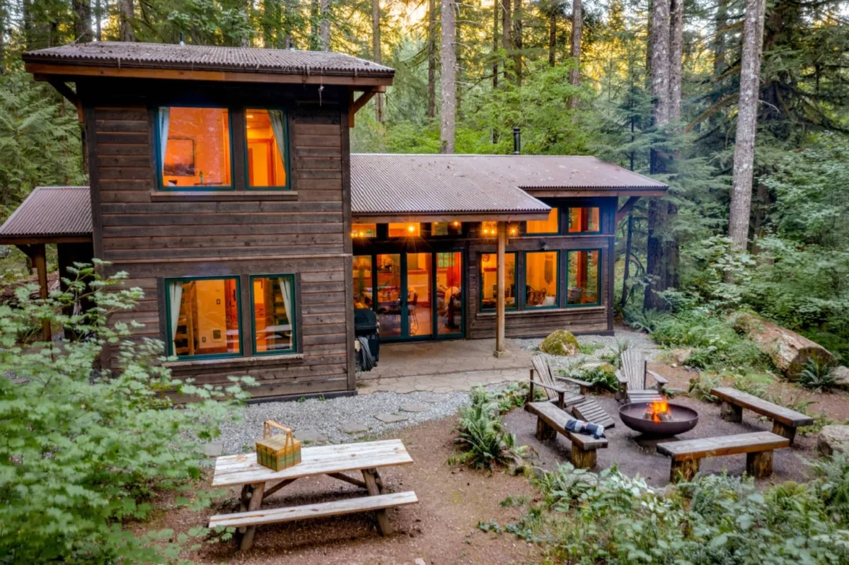 This exterior shot displays the wooden cabin nestled among tall trees, with a fire pit surrounded by chairs and a picnic table set up nearby. The lush greenery and open outdoor space showcase areas where families can gather, play, or relax, evoking a sense of adventure and shared experiences.