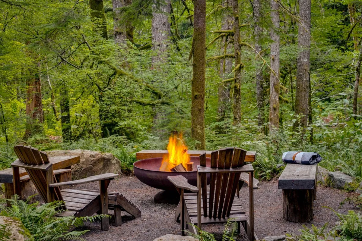 A cozy fire pit area with wooden chairs and stone seating, set beneath a canopy of trees. A blanket on the bench adds a touch of warmth, while the fire pit glows invitingly, evoking communal gatherings and storytelling under the stars.