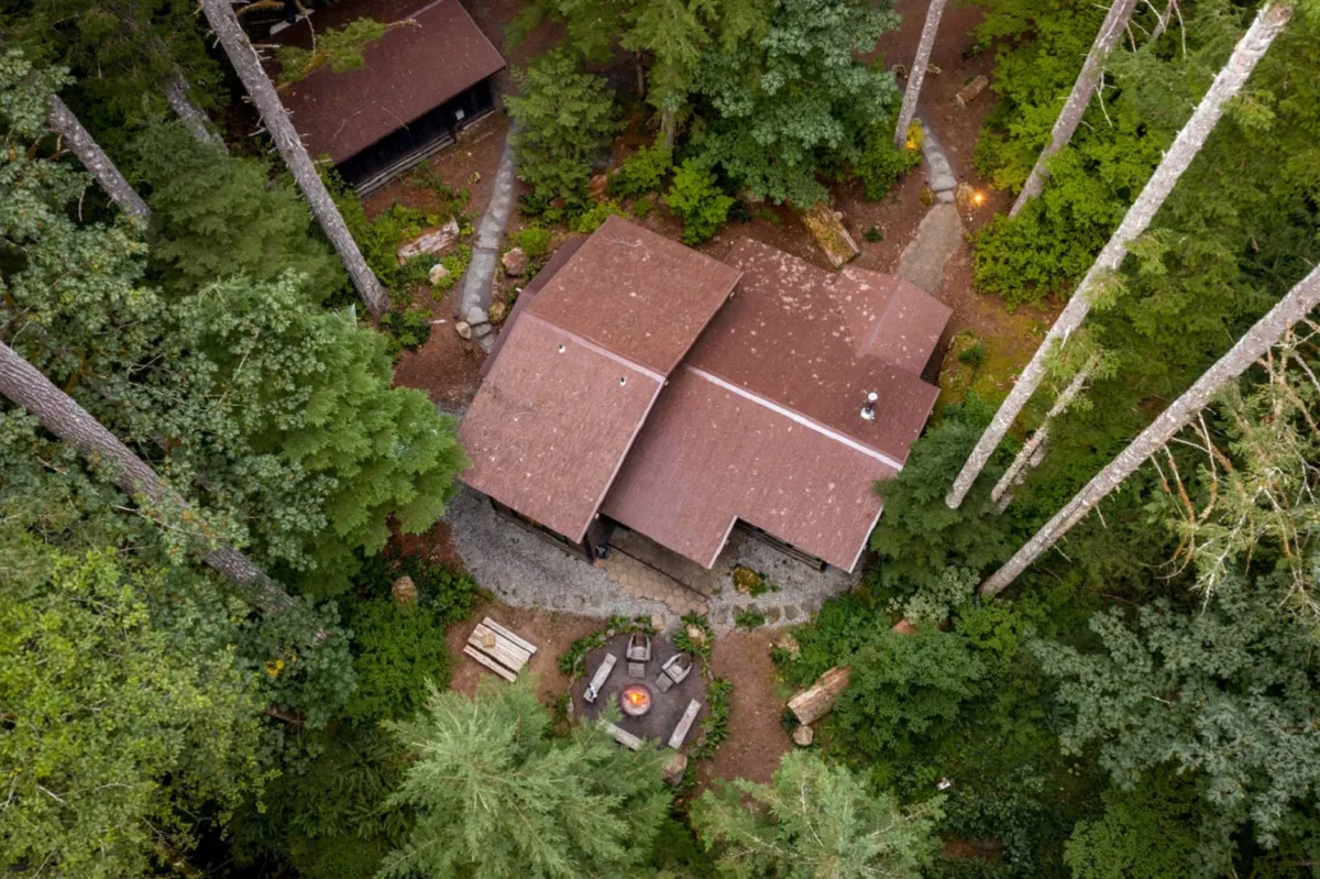 An exterior view of the main house nestled among trees, featuring a fire pit and expansive deck area. The scene invites outdoor relaxation, nature walks, and evenings spent under the stars.