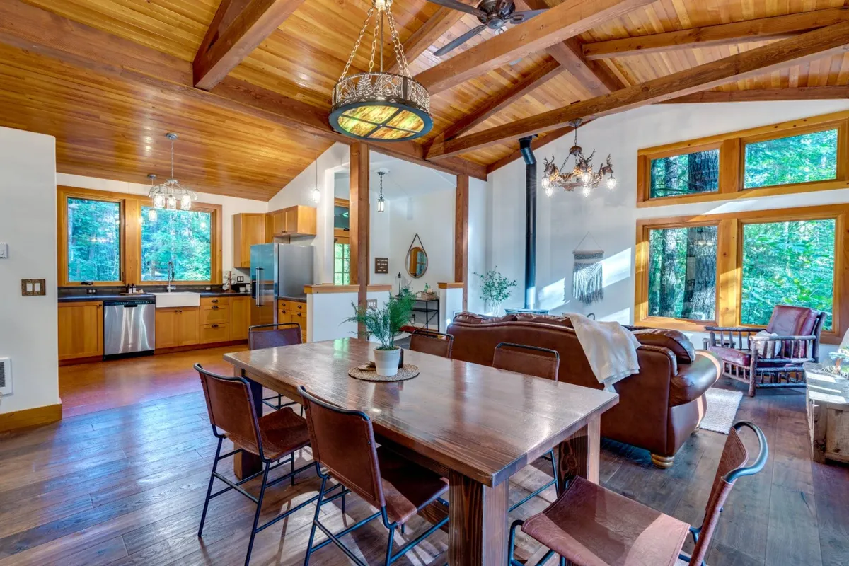An open-concept space with a wooden dining table set near large windows, a plush sofa, and a visible kitchen area in the background. The arrangement encourages conversation and shared activities, all bathed in natural light with views of the surrounding forest.
