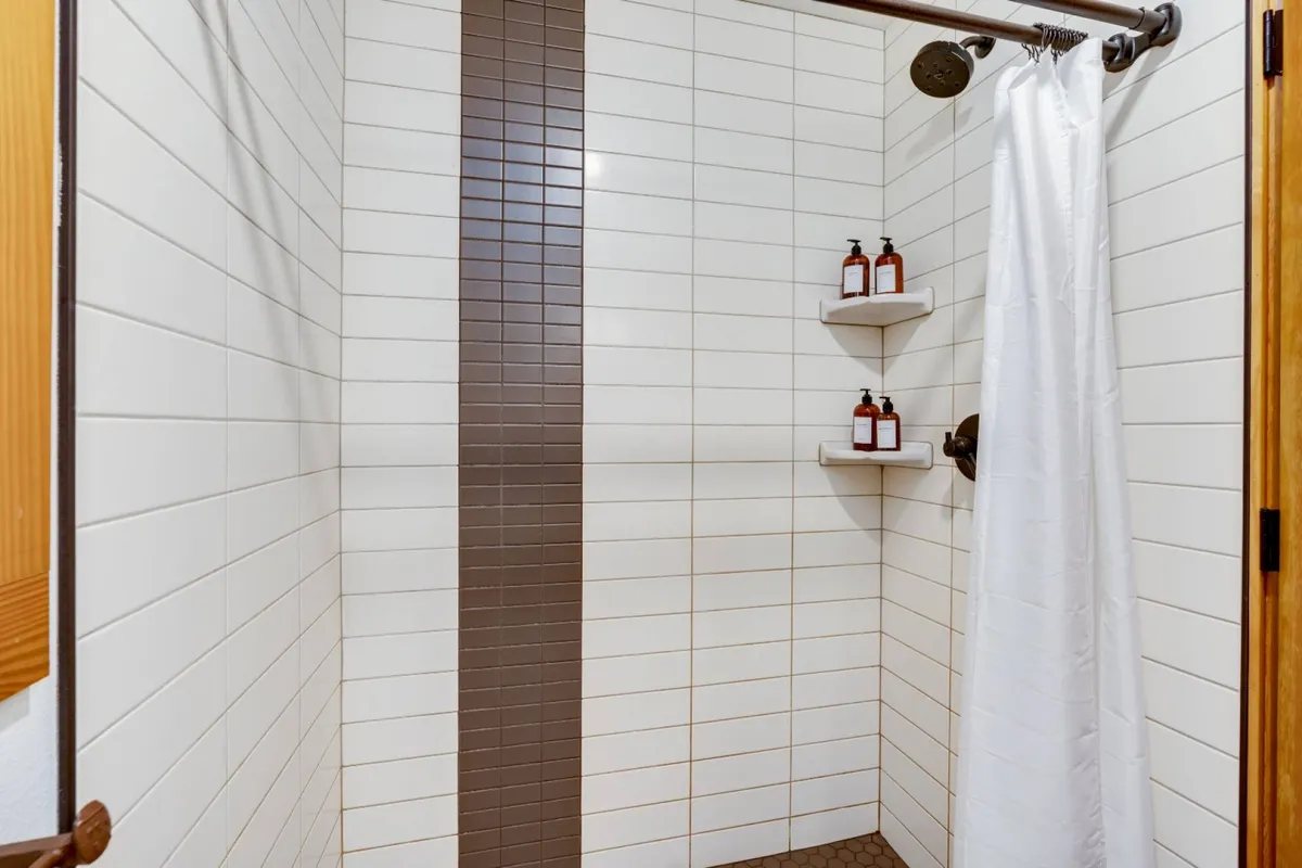 A soothing, spa-like walk-in shower with a modern showerhead, shelves with toiletries, and a white curtain. The atmosphere is clean and calming, inviting guests to refresh after a day of adventure.