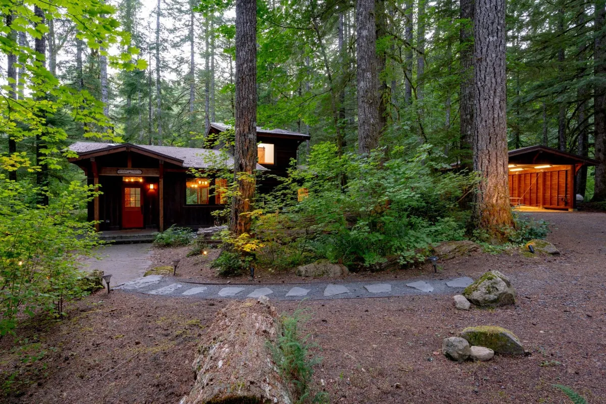 This exterior image captures the wooden cabin nestled among towering trees, with a gently curving pathway inviting guests to wander into the forest. The landscaping merges seamlessly with the natural woods, providing a glimpse of the quiet, immersive woodland setting around Tamanawas Cabin.