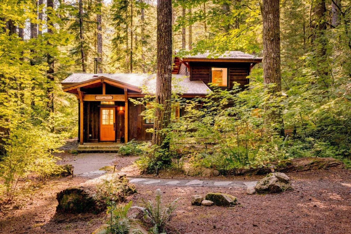 The wooden cabin sits quietly among towering trees, with a stone pathway leading to its porch. The setting is serene, suggesting peaceful mornings and calm evenings surrounded by nature.