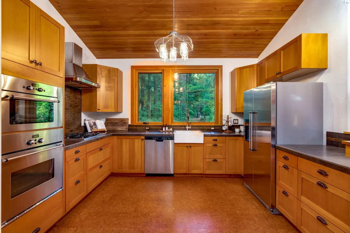 A modern kitchen boasting a central island sink, stainless steel appliances, and wooden cabinetry, with expansive windows showcasing a vibrant forest view. The layout is perfect for cooking, gathering, and enjoying sunlight or forest vistas while prepping meals.