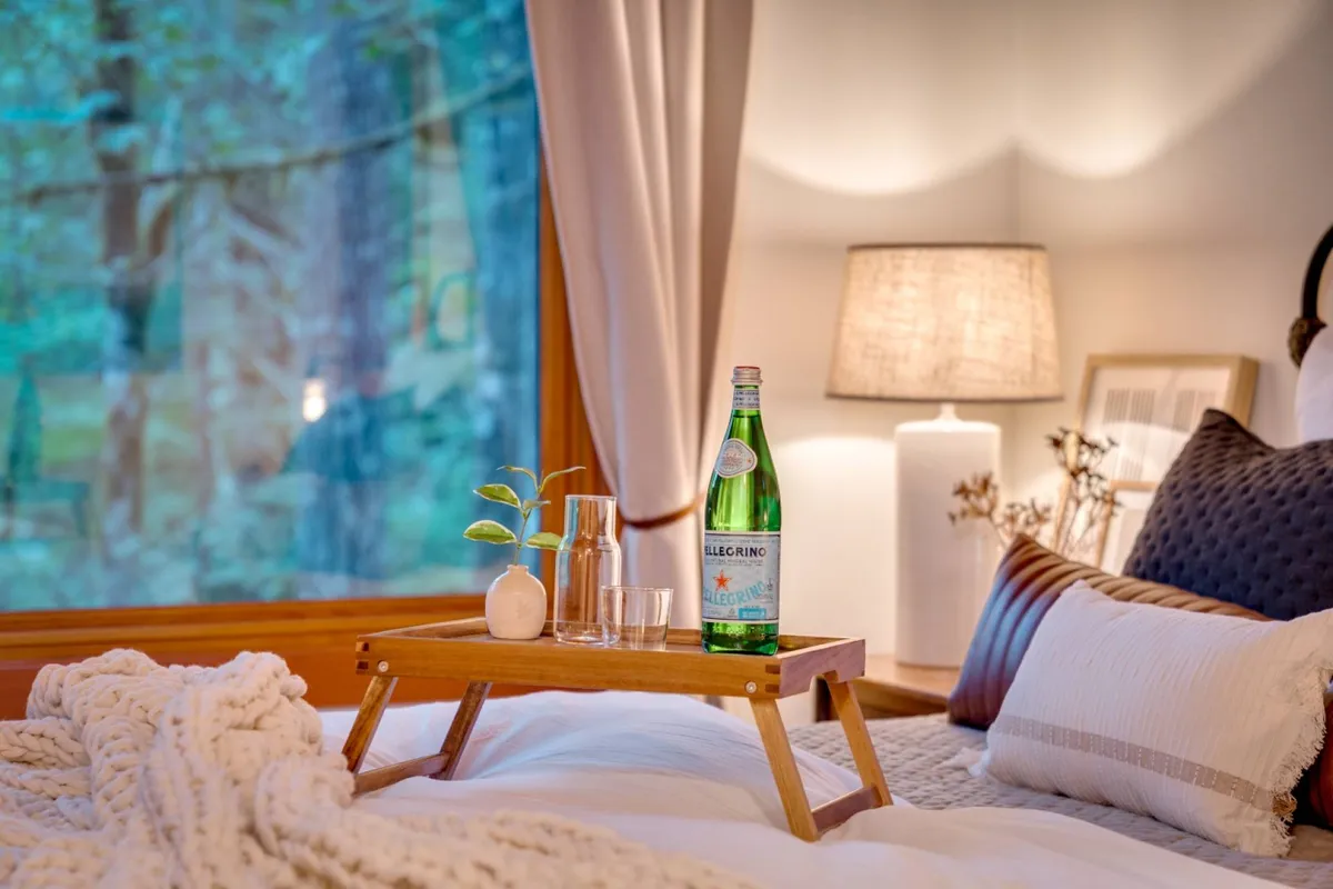 A tranquil bedroom featuring a cozy bed with a soft blanket, a bedside glass bottle of sparkling water, a decorative lamp, and a window framing serene views of the outdoors. The overall mood is restful and inviting, perfect for unwinding after a day outside.