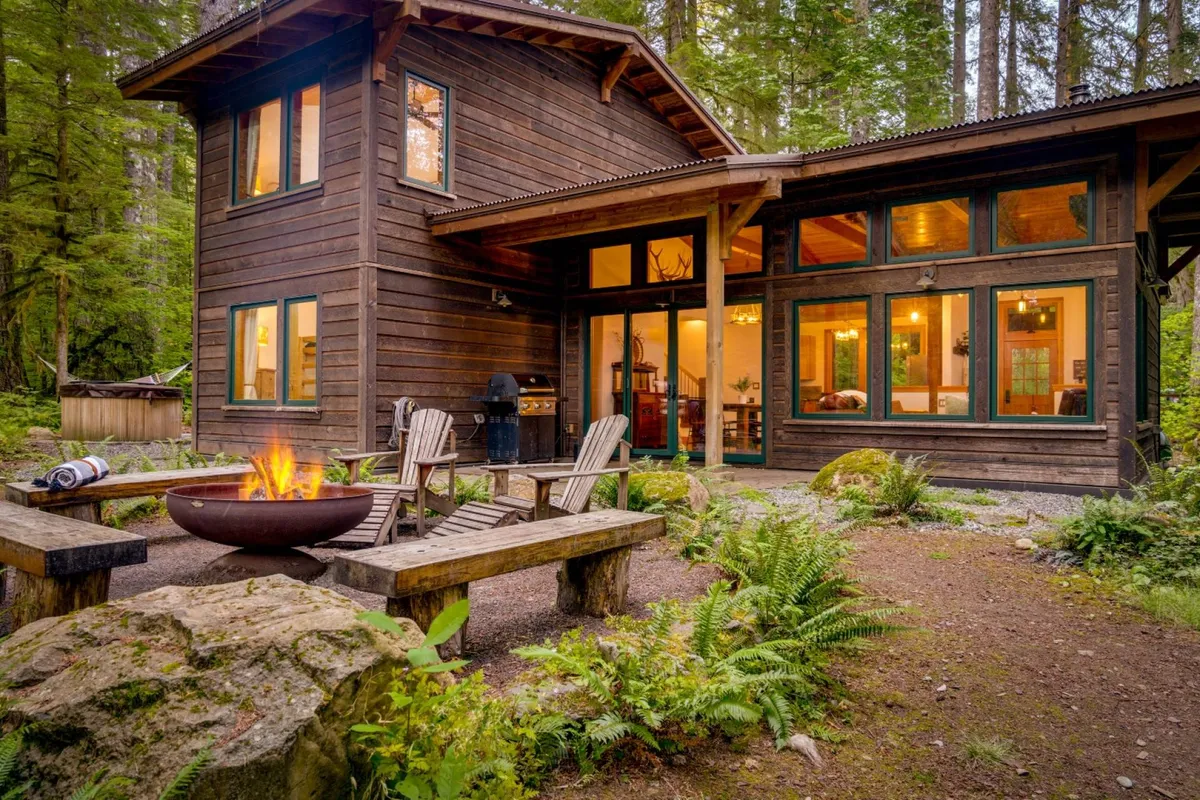 An exterior shot featuring a fire pit surrounded by wooden chairs set amidst tall trees. The large windows of the cabin glow warmly in the background, inviting guests to gather and enjoy the tranquil forest setting under the open sky.