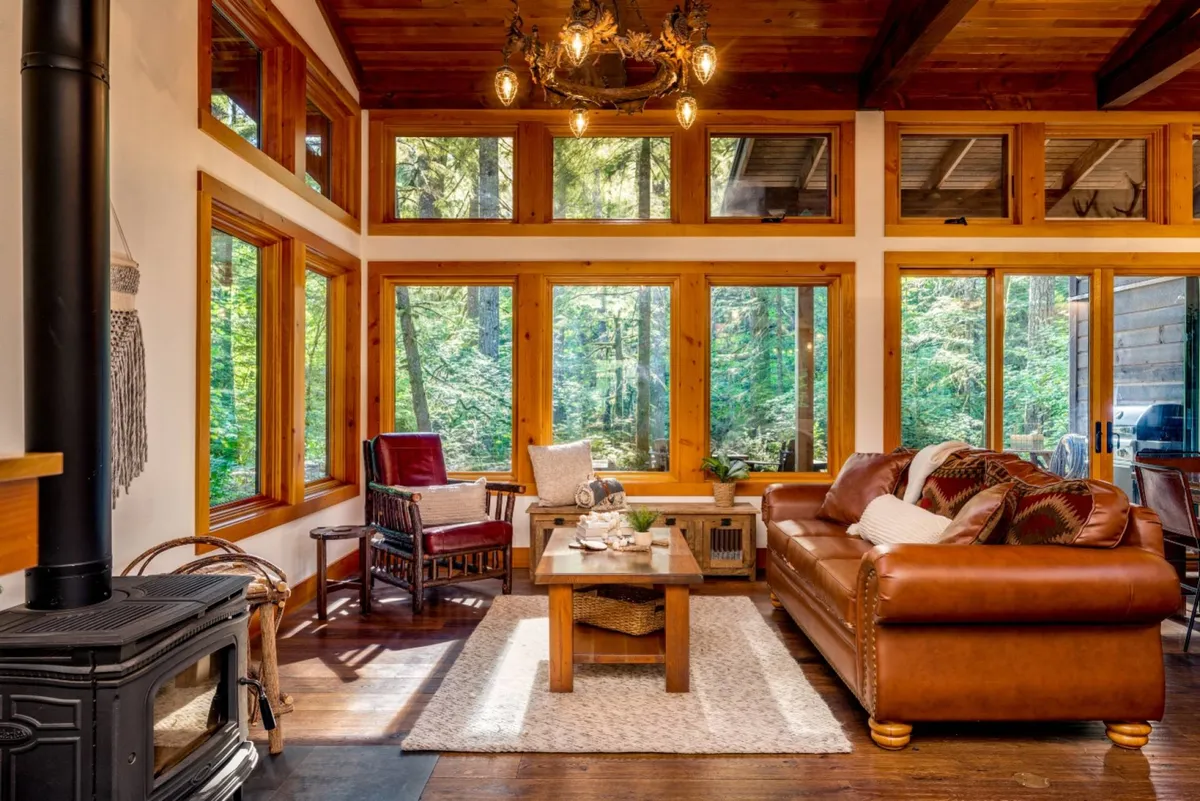 A spacious living room featuring a leather sofa, a sturdy wooden coffee table, and a classic wood-burning stove. Large windows frame lush forest views, flooding the space with natural light and offering a seamless visual connection to the outdoors. The atmosphere is warm and welcoming, ideal for family gatherings or relaxing evenings by the fire.