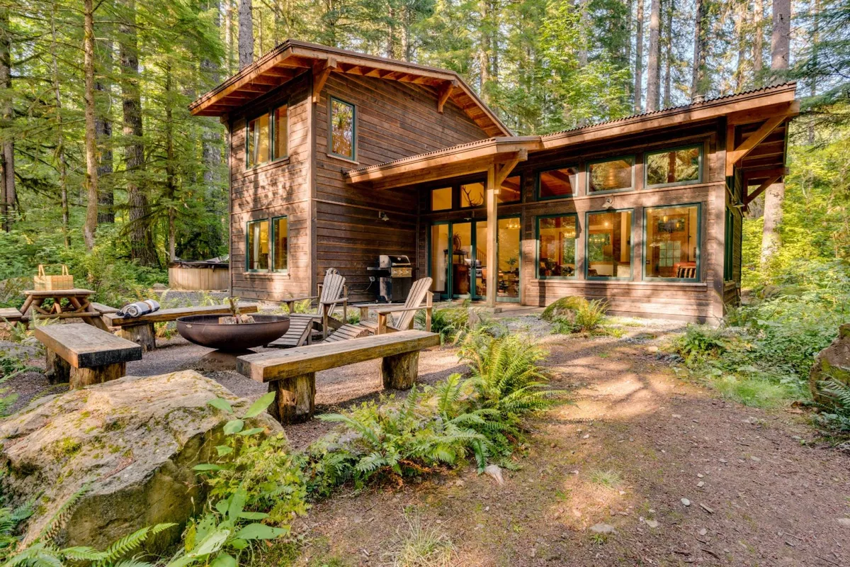 A modern wooden house with an inviting outdoor seating area and a fire pit, all set amid a lush forest. The image captures both the covered patio and fire pit in the same frame, with the surrounding nature enhancing the sense of retreat.